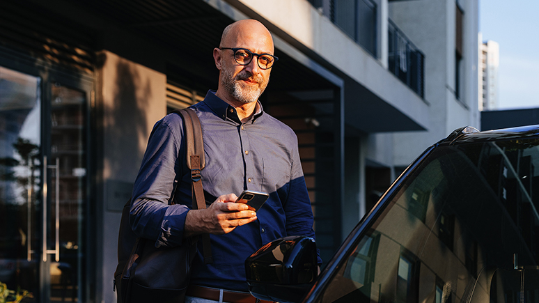 a man standing by a car, holding a phone and a bag