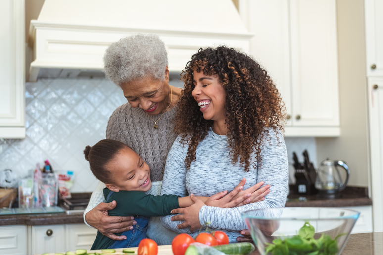 A beautiful senior woman of African descent embraces her granddaughter and daughter in a loving hug. They are bonding in the kitchen one afternoon. There is a lot of natural light.