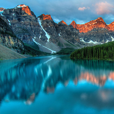 Sunset illuminating mountains near reflective lake and green pine tree forest