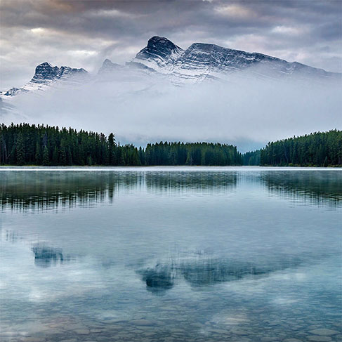 Snow-capped mountain covered in fog near body of water and trees
