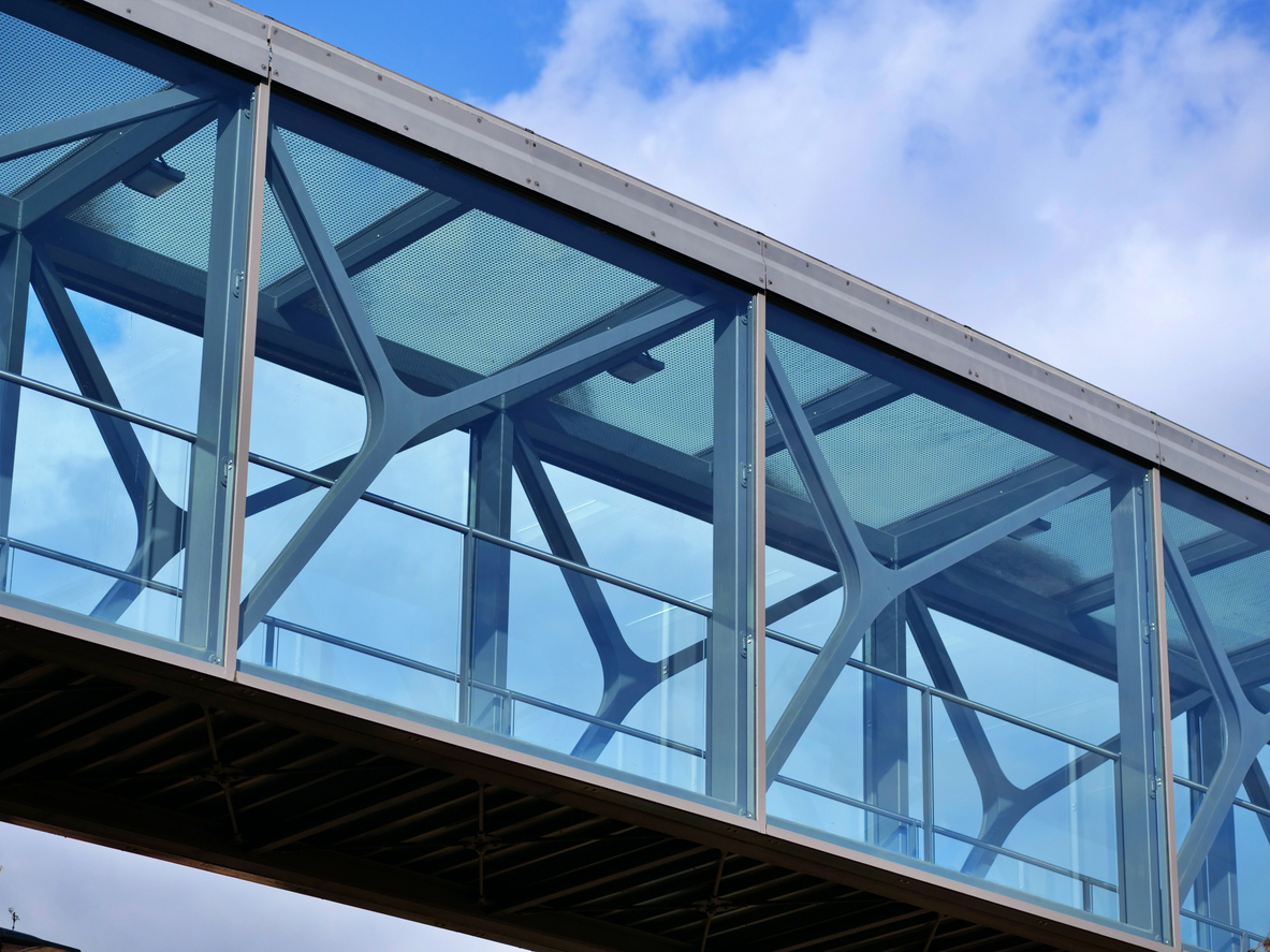 Low-angle view of a modern glass skybridge featuring a distinctive bionic steel truss structure. The intricate, organic metal framework is visible through transparent blue-tinted glass panels, reflecting a bright sky with soft white clouds. The architectural design combines industrial strength with geometric elegance, creating a sleek, futuristic aesthetic in an urban environment.