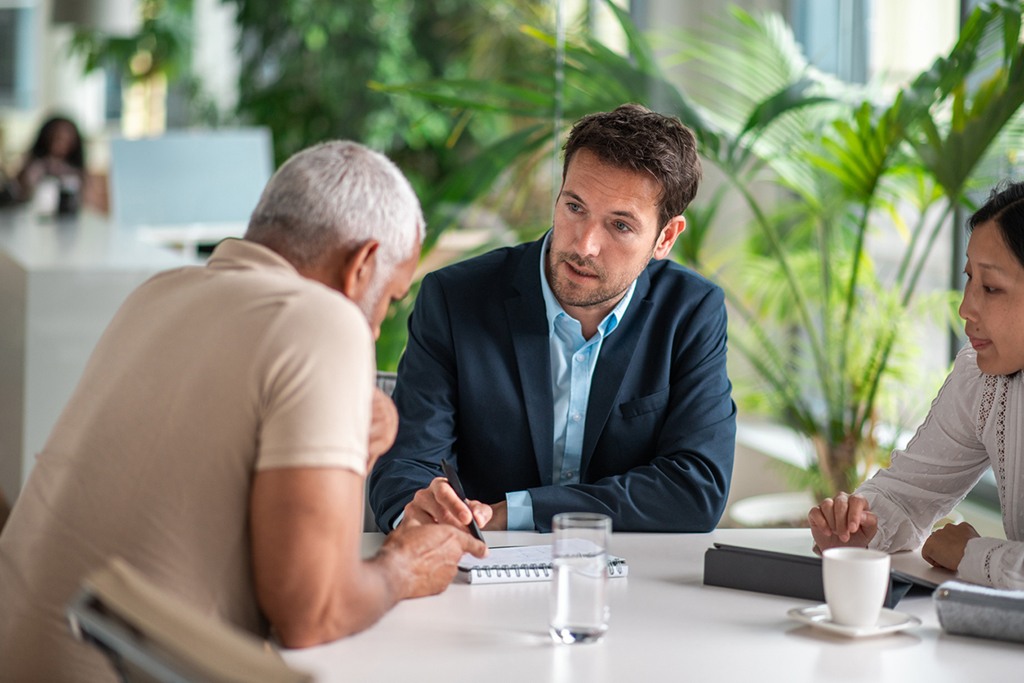 A senior Hispanic man consults with Asian and Caucasian financial advisors in a bright office, discussing strategies with focus and professionalism.