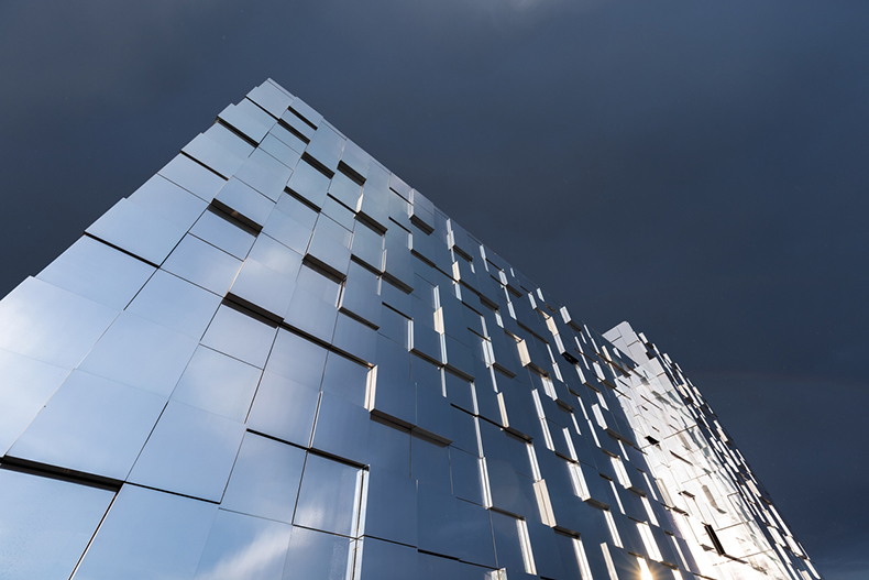 Low-angle view of a modern reflective building facade with mirrored glass panels and geometric patterning against a dramatic stormy sky, highlighting contemporary architecture and urban design