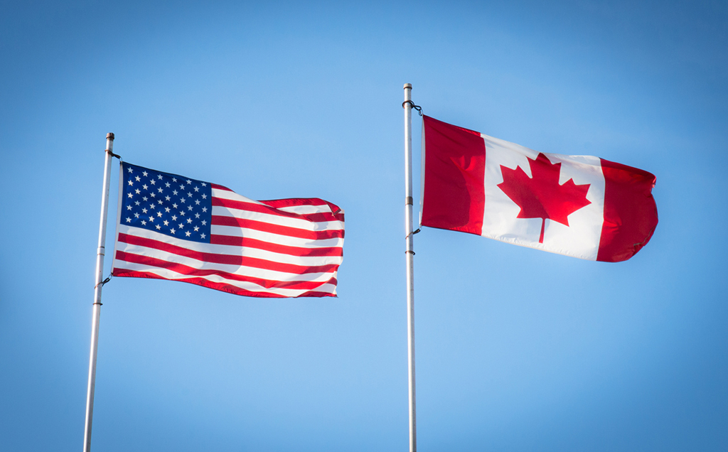 Two flags in front of the blue sky. An american flag on the left. a canadian flag on the right